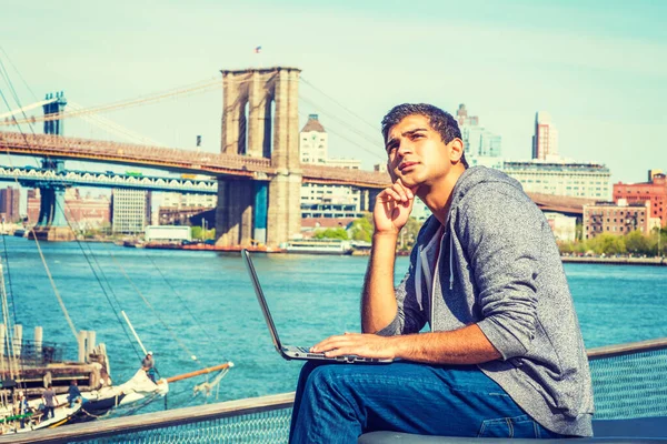 East Indian American student traveling, studying in New York, sitting by river, working on laptop computer, tilting head, hand touching cheek, looking up, thinking. Brooklyn bridge on background.