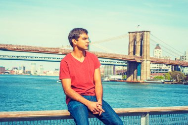 Seni özledim, seni bekliyorum. Doğu Hint asıllı, V yaka tişörtlü, kot pantolonlu, nehir kenarındaki çitlerde oturan ve etrafa bakıp düşünen bir adam. Manhattan ve Brooklyn köprüleri arka planda.