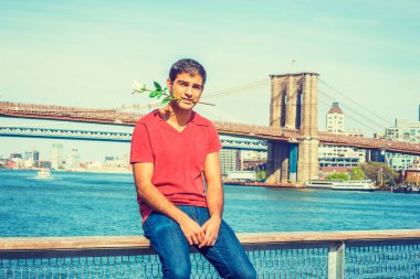 Genç yakışıklı adam seni bekliyor. Doğu Hint asıllı, V yaka tişörtlü, kot pantolonlu, nehir kenarına oturmuş, elinde beyaz bir gül tutuyor. Manhattan ve Brooklyn köprüleri arka planda.