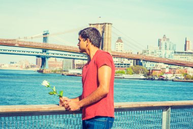 Seni özlüyorum, seni bekliyorum. Doğu Hint asıllı, kırmızı tişört giyen, nehrin kenarında duran, elinde beyaz bir gül tutan, başka tarafa bakan bir adam. Manhattan ve Brooklyn köprüleri arka planda. 
