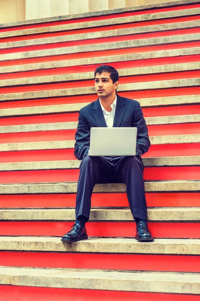 Way to Success. East Indian American College Student studies in New York. Wearing black suit, white shirt, leather shoes, young man sits on stairs, works on laptop computer, thinks.