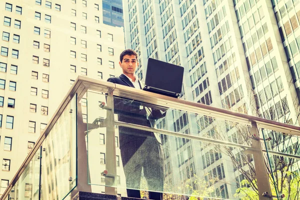 Way to Success. East Indian American student studies in New York. Wearing black suit, young man stands by railing in business district, works on laptop computer, looks away, thinks.