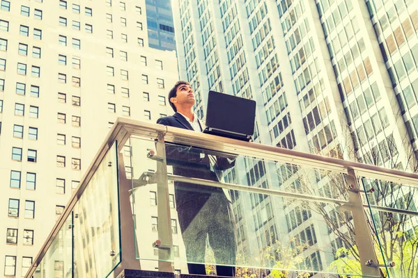 Way to Success. East Indian American student studies in New York. Wearing black suit, young man stands by railing in business district, works on laptop computer, looks up, thinks.