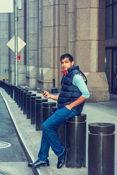 Modern City Life. East Indian American College Student wearing long sleeve T shirt, blue down vest, jeans, leather shoes, sitting on pillar on vintage street in New York, texting.