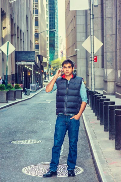 Modern City Life. East Indian American Man wearing long sleeve T shirt, blue down vest, jeans, leather shoes, stands on narrow vintage street in New York, talks on cell phone.
