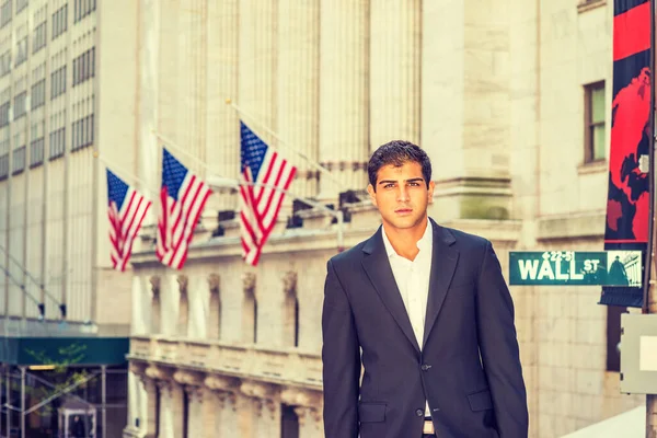 East Indian American Business Man travels, works in New York. Wearing black suit, a college student stands on Wall Street with vintage buildings with American flags, looking at you.