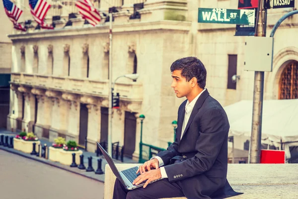 East Indian American Business Man travels, works in New York. Wearing black suit, college student sits on Wall Street by vintage office building with flags, works on laptop computer.