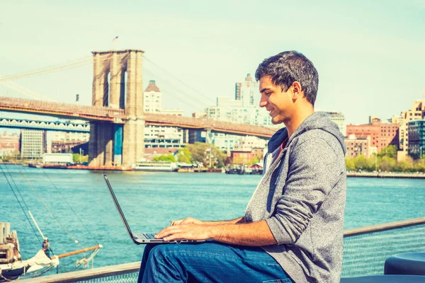 Başarının yolu. Mutlu Doğu Hintli Amerikalı üniversite öğrencileri New York 'ta okuyor, kapüşonlu sweatshirt giyiyor, kot pantolon giyiyor, nehir kenarında oturuyor, dizüstü bilgisayarla çalışıyor. Brooklyn Köprüsü arka planda.