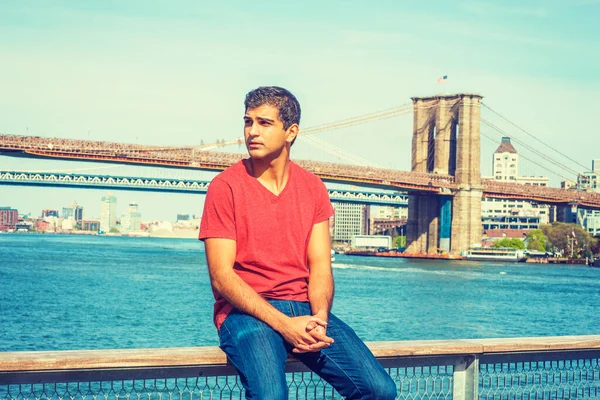 I miss you, waiting for you. East Indian American man wearing red V neck T shirt, jeans, sits on fence by river, looks around, thinks. Manhattan, Brooklyn bridges on background.