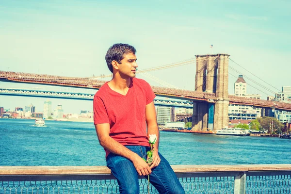 I miss you. Young East Indian American man wearing red V neck T shirt, jeans, sits on fence by river, holds white rose, looks around, thinks. Manhattan, Brooklyn bridges on background.