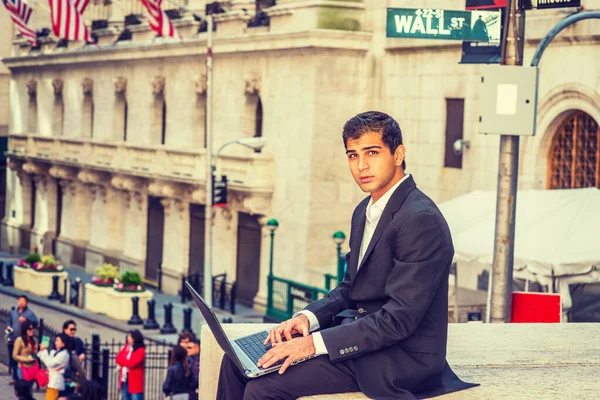East Indian American Business Man travels, works in New York. Wearing black suit, college student sits on Wall Street by vintage office building, works on laptop computer.