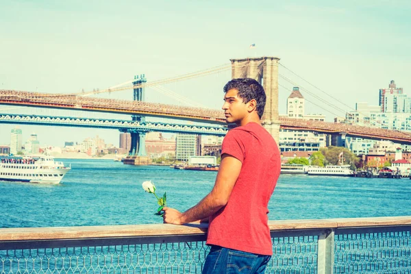 Seni özlüyorum, seni bekliyorum. Doğu Hint asıllı, kırmızı tişört giyen, nehrin kenarında duran, elinde beyaz bir gül tutan, başka tarafa bakan bir adam. Manhattan, Brooklyn köprüleri, arka planda tekneler. 