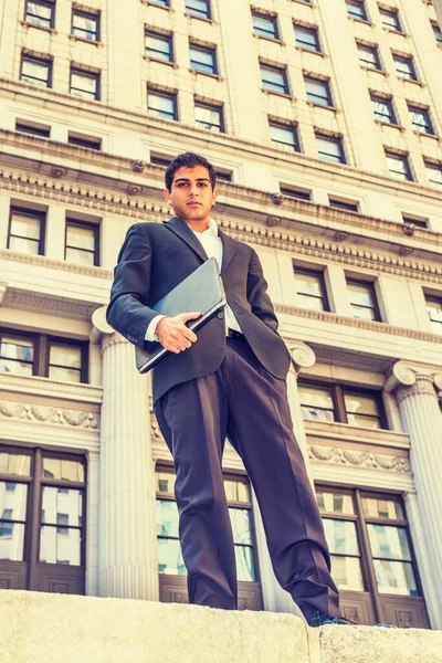 East Indian American college student travels, studies in New York. Wearing black suit, white shirt, holding laptop computer, young man stands outside vintage office building, looking forward.