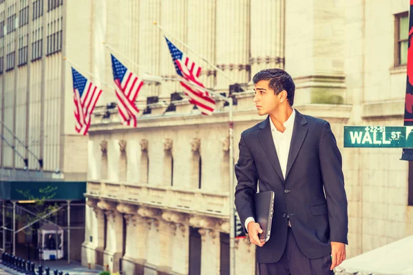 East Indian American Business Man travels, works in New York. Wearing black suit, holding laptop computer, college student stands on Wall Street with American flags, looks away.