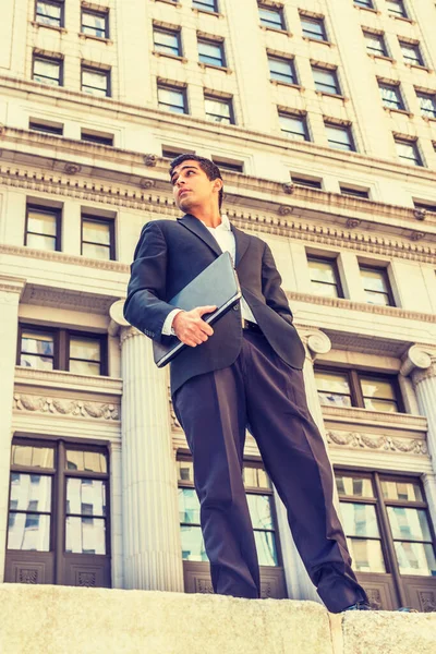 East Indian American college student travels, studies in New York. Wearing black suit, white shirt, holding laptop computer, young man stands outside vintage office building, looking forward.