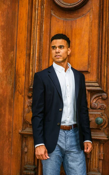 African American Businessman working in New York. Wearing black blazer, white shirt, gray pants, a college student standing by vintage office doorway, looking up, thinking.