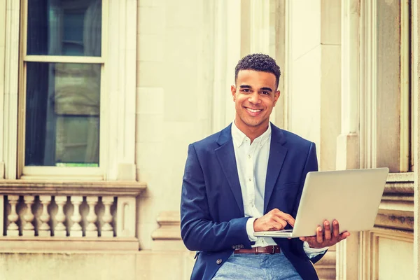 African American college student studying in New York, wearing black blazer, sitting inside vintage office building on campus, reading, working on laptop computer, smiling.