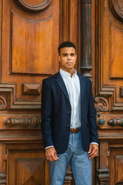 African American Businessman working in New York. Wearing black blazer, white shirt, gray pants, black college student standing by vintage office doorway on campus, seriously looking at you, thinking.