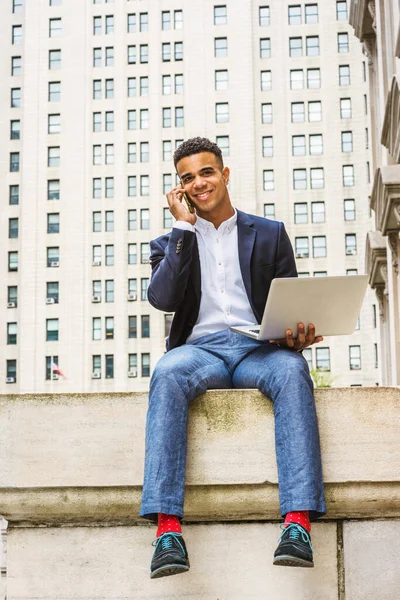 New York 'ta okuyan Afro-Amerikan üniversite öğrencisi, mavi ceket, gri pantolon, beyaz gömlek, kırmızı çorap, spor ayakkabılar, sokakta ofis dışında oturan, dizüstü bilgisayarla çalışan, telefonla konuşan