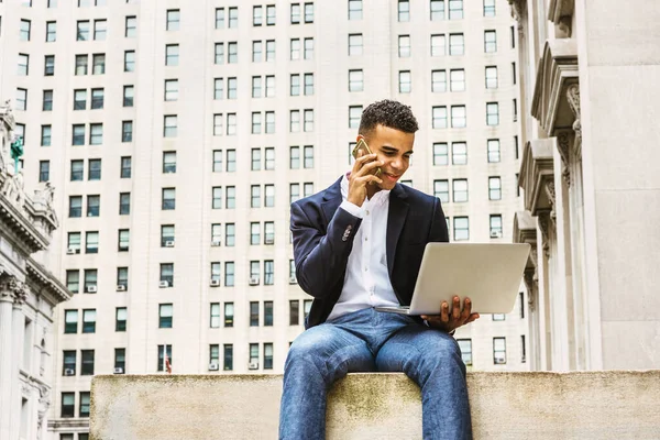 Modern life. African American college student studying in New York, sitting by vintage office building on street, reading, working on laptop computer, talking on cell phone.