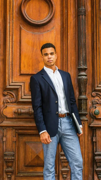 African American Businessman working in New York. Wearing black blazer, white shirt, gray pants, holding laptop computer, a college student standing by vintage office doorway on campus, looking at you