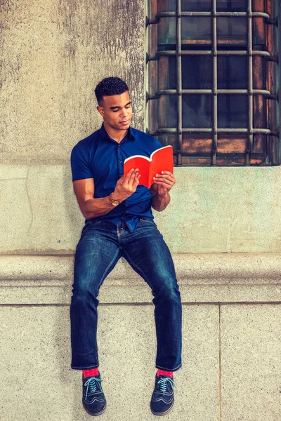 African American College Student studying in New York, wearing blue short sleeve shirt, jeans, red socks, sneakers, sitting against wall on street, reading red book.