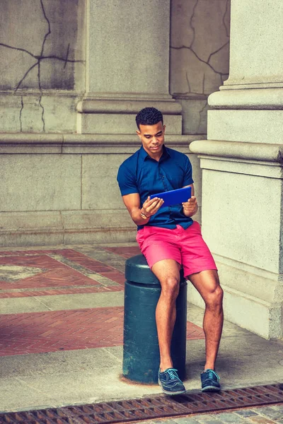 Şehirde yaz. Mavi kısa kollu gömleği, kırmızı şortu, spor ayakkabıları, New York 'ta metal sütunda oturan Afro-Amerikan üniversite öğrencisi, küçük tablet bilgisayar okuyor.