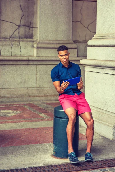 Summer in City. Modern life. Wearing blue short sleeve shirt, red shorts, sneakers, African American college student sitting on metal pillar on street in New York, reading small tablet computer.