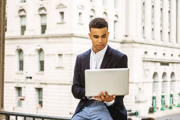 African American college student studying in New York, wearing black blazer, sitting on railing by vintage office building on campus, reading, working on laptop computer.