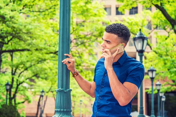 African American college student calling outside, wearing blue short sleeve shirt, standing by light pole on street on campus in New York in summer, tilting head, listening, talking on cell phone