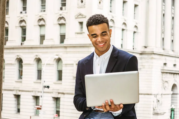 Happy African American college student studying in New York, wearing black blazer, sitting by vintage office building on campus, reading, working on laptop computer, smiling.