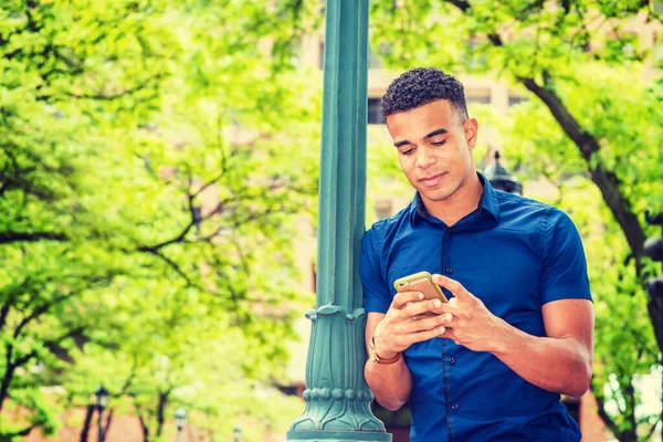 Afro-Amerikan üniversite öğrencisi dışarıda mesajlaşıyor, mavi kısa kollu tişört giyiyor, New York 'ta sokak lambasına yaslanmış, cep telefonundan mesajlar okuyor.. 