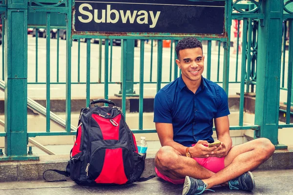 African American college student traveling, studying in New York, wearing blue short sleeve shirt, red shorts, sneakers, bag with bottle water on ground, sitting on street by Subway sign, texting