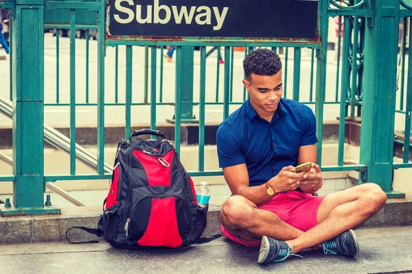 African American college student traveling, studying in New York, wearing blue short sleeve shirt, red shorts, sneakers, bag with bottle water on ground, sitting on street by Subway sign, texting