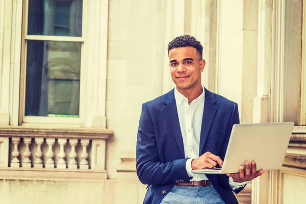 African American college student studying in New York, wearing black blazer, sitting inside vintage office building on campus, reading, working on laptop computer, smiling.