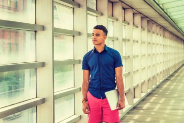 Okulda yaz. Kısa kollu gömleği, kırmızı şortu, dizüstü bilgisayarı olan, Afrika kökenli Amerikalı bir öğrenci cam duvarlı, tavanlı, New York kampüsünün ahşap zemininde duruyor.