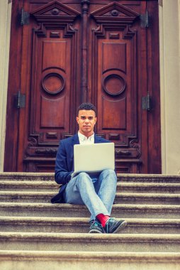 Başarının yolu. Mavi ceket, pantolon, kırmızı çorap, spor ayakkabı, bacak bacak üstüne atan Afro-Amerikan üniversite öğrencisi New York 'ta kampüste klasik kapının önünde oturuyor, dizüstü bilgisayarla çalışıyor..