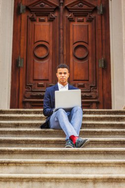 Başarının yolu. Mavi ceket, pantolon, kırmızı çorap, spor ayakkabı, bacak bacak üstüne atan Afro-Amerikan üniversite öğrencisi New York 'ta kampüste klasik kapının önünde oturuyor, dizüstü bilgisayarla çalışıyor..