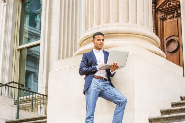 Başarının yolu. Afrikalı Amerikalı üniversite öğrencisi New York 'ta okuyor. Mavi ceketli, beyaz gömlekli, gri pantolonlu genç adam ofis binasının dışındaki sütuna karşı duruyor, dizüstü bilgisayarla çalışıyor..