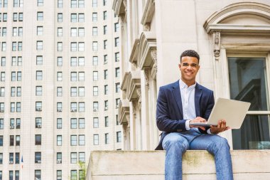 New York 'ta Afro-Amerikan öğrencilerin mutlu çalışmalar. Mavi ceketli, beyaz gömlekli, kampüsteki klasik ofis binasının önünde oturan, dizüstü bilgisayarında çalışan, gülümseyen genç bir zenci.