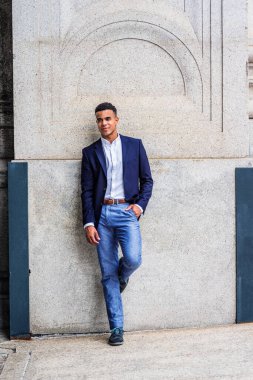 New York 'ta Afro-Amerikan bir adamın portresi. Mavi ceketli, beyaz gömlekli, pantolonlu, spor ayakkabılı, erkek bir üniversite öğrencisi kampüsteki klasik duvarın karşısında duruyor.. 