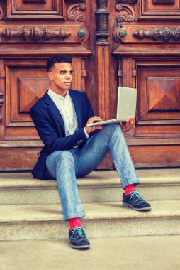 Başarının yolu. Mavi ceketli, gri pantolonlu, spor ayakkabılı, Afrikalı Amerikalı üniversite öğrencisi New York 'ta kampüsün klasik ofis kapısında oturuyor, dizüstü bilgisayarla çalışıyor.. 