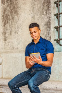 Okumayı seviyorum. Modern okuma. Mavi kısa kollu gömlek, kot pantolon, Afro-Amerikan üniversite öğrencisi New York 'ta eski moda bir duvarın karşısında oturmuş, küçük tablet bilgisayar okuyor..