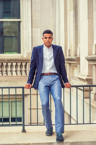 Mavi alevli, beyaz gömlekli, gri pantolonlu, spor ayakkabılı, Afro-Amerikan erkek üniversite öğrencisi New York 'ta kampüste eski moda ofis binasında korkuluklarda oturuyor..