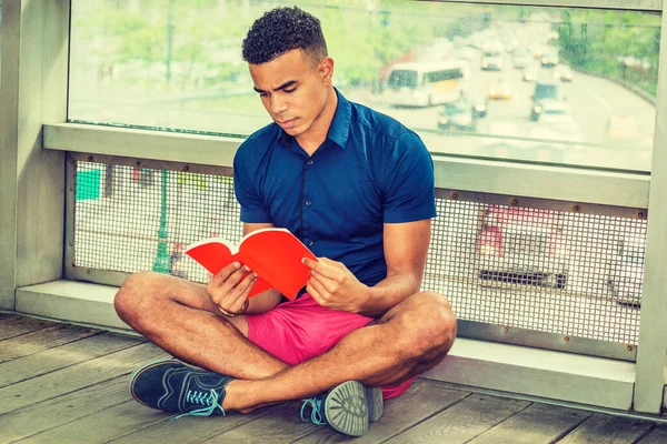 Way to Success. I Love Reading Book. African American college student sitting cross legged on floor against glass wall on campus, looking down, reading red book. Street, many cars on background.