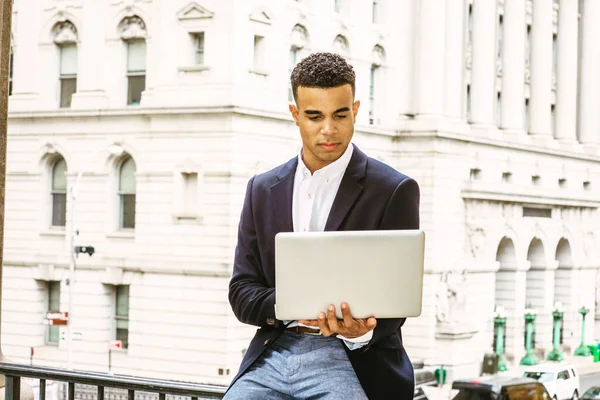 African American college student studies in New York.  Wearing black blazer, young guy sits on railing by vintage office building on campus, reads, works on laptop computer.