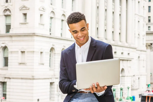 African American college student studies in New York. Wearing black blazer, young happy guy sits on street by vintage office building on campus, reads, works on laptop computer.