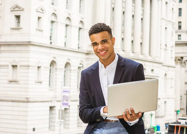 African American college student studies in New York. Wearing black blazer, young happy guy sits on street by vintage office building on campus, reads, works on laptop computer.