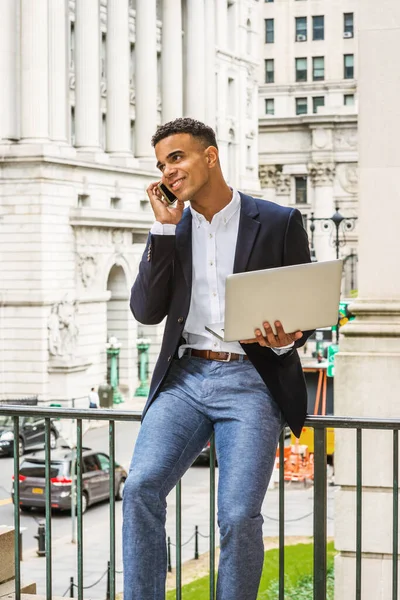 African American Businessman works in New York. Wearing black blazer, white shirt, young man sits on street by vintage office building, works on laptop computer, talks on cell phone.