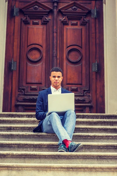 Başarının yolu. Mavi ceket, pantolon, kırmızı çorap, spor ayakkabı, bacak bacak üstüne atan Afro-Amerikan üniversite öğrencisi New York 'ta kampüste klasik kapının önünde oturuyor, dizüstü bilgisayarla çalışıyor..